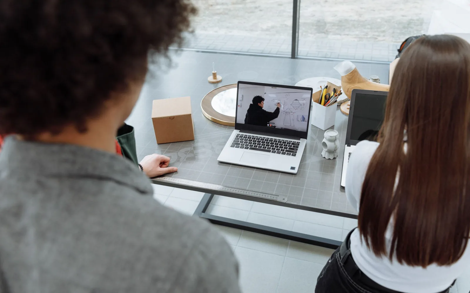 Twee collega's achter een laptop tijdens een werksessie, één wijst naar schetsen op een whiteboard.