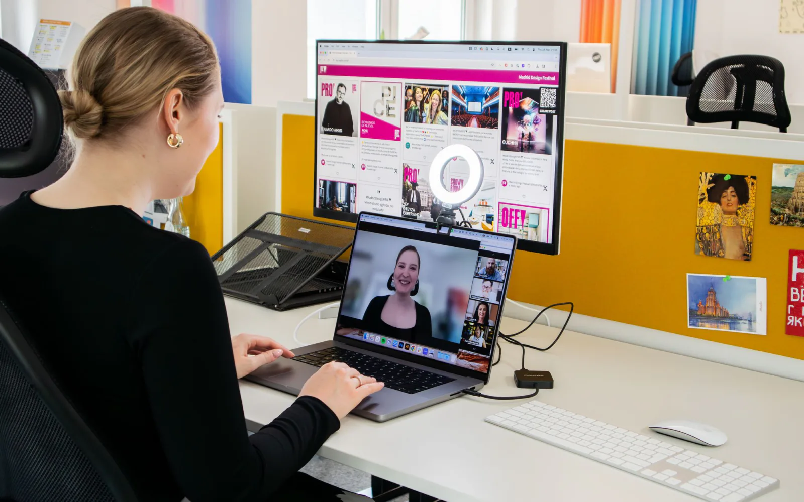 Vrouw aan haar bureau met een laptop in videogesprek met vijf collega's en een extern scherm met een Spaans design-dashboard.