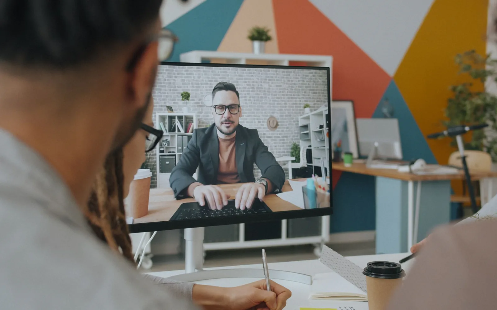 Over-de-schouder opname van een deelnemer in een hybride videogesprek in een kantoor met warme kleurenaccenten.