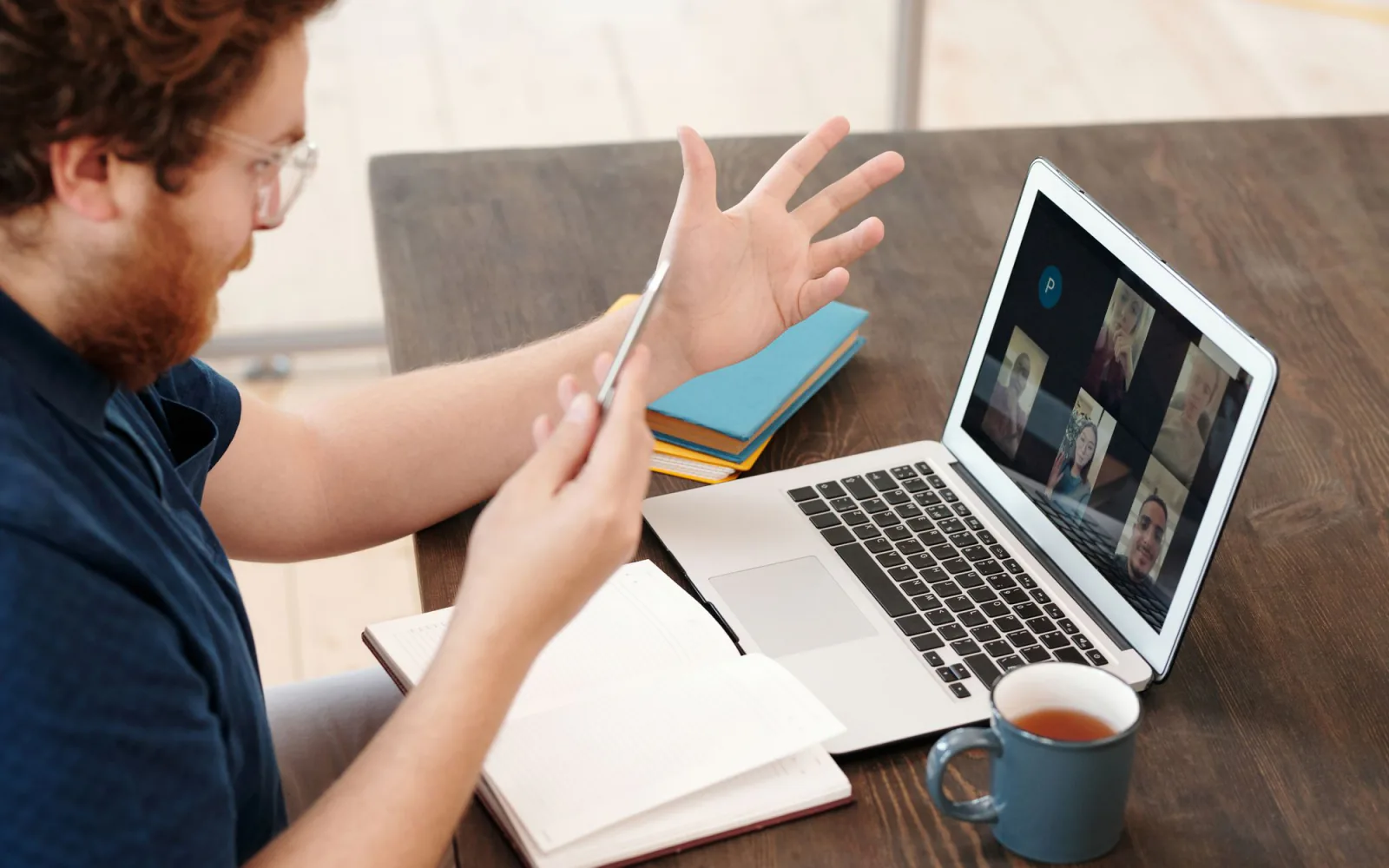 Man in een thuiskantoor met een laptop; hij gebaart naar het videogesprek-grid op het scherm, mok thee ernaast.